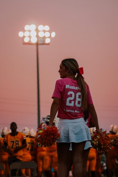 On September 8th, 2023, The Springfield community mourned the loss of a beloved teacher at Kickapoo High School, Ms. Barron. She was cheer coach who was loved by many and was described by many people as a bright light in everyones life. At that Fridays football game, the cheer squad organized a balloon release in her honor. As expected, the stand were packed out with supporters. Our Cheer and Poms teams wore pink matching t-shirts that said Momma B on the back and Be The Sunshine on the front, which was a saying she constantly said to everyone in her life. Senior Joslyn Magers, a cheer team member who was very close to Ms. Barron, proudly shows off her shirt to the crowd. 